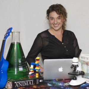 Jen Tuttle Parsons sits smiling with a laptop, a microscope, books, LEGOs, and an Erlenmeyer flask