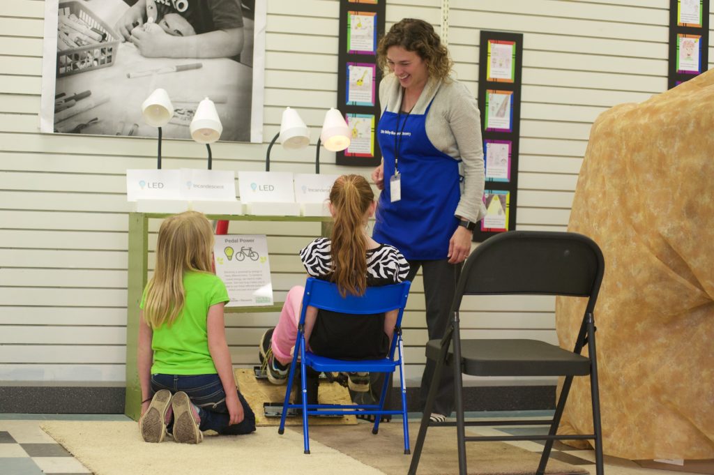 Jen, in blue apron, speaks to two young museum guests while demonstrating an exhibit comparing LED, incandescent, and fluorescent light bulbs.